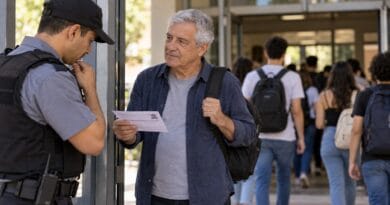 Homem mais velho mostra o comprovante de inscrição a um segurança na entrada de um local de vestibular, enquanto candidatos mais jovens entram ao fundo.